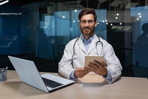 retrato de un joven hermoso masculino médico quien se sienta y trabajos en un oficina en un hospital a un mesa con un ordenador portátil. sostiene un tableta en su manos, mira a el cámara, sonrisas foto