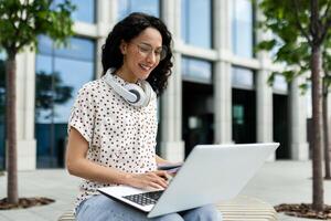 Smiling young woman with headphones using laptop while sitting on a bench outside, surrounded by modern urban architecture. photo