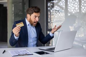 enojado joven hombre en un traje trabajando en el oficina, sentado a un escritorio, participación un crédito tarjeta, mirando preocupadamente a el ordenador portátil pantalla, lanzamiento arriba su manos en frustración. foto