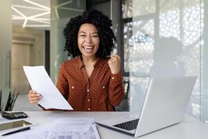 retrato de exitoso africano americano mujer jefe a lugar de trabajo, mujer de negocios detrás papel trabajo sonriente y mirando a cámara, participación mano arriba ganador y triunfante gesto dentro oficina. foto