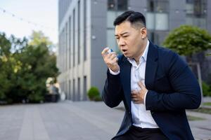 A young Asian man suffering from cough and asthma in a business suit is sitting near an office building, holding his hands to his chest and using an inhaler. photo