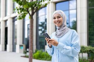 joven hermosa mujer de negocios en hijab camina en el ciudad desde fuera de el oficina edificio, mujer usos un solicitud en el teléfono, sonrisas y mira a el cámara, sostiene un teléfono inteligente para en línea llamar. foto