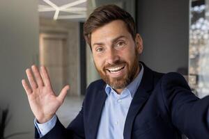 de cerca retrato de un sonriente y exitoso joven empresario en un traje en pie en el oficina, ondulación y saludo con su mano en el cámara teléfono, hablando en llamar. foto