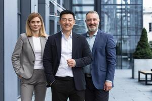 Successful diverse business team, three workers smiling and looking at camera, dream team with asian boss outside office building, colleagues in business suits photo