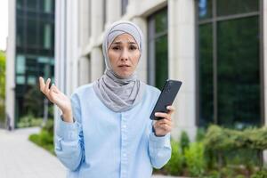 trastornado islámico mujer extensión manos en Confusión mientras participación teléfono inteligente en urbano antecedentes. estresado dama en hijab y azul camisa luchando con utilizando nuevo digital artilugio y mirando para ayuda. foto