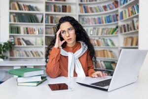 irritado público biblioteca trabajador laminación ojos y propensión cabeza en mano por escritorio con computadora y libros. irritado mujer sensación enojado acerca de ruidoso comportamiento en espacio de trabajo con insatisfecho cara expresión. foto