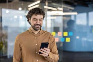 Carefree guy with stylish hairstyle using black cell phone while standing alone in working space. Relaxed man watching interesting in social media while using wireless internet connection. photo