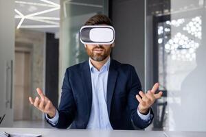 A young man sits in the office at the table in front of the camera in a virtual online mask, talks remotely, conducts a meeting, gestures with his hands. photo