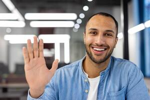 Close-up portrait of a young hispanic male office worker and freelancer sitting in front of the camera, greeting and waving at the camera. photo