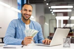 Portrait of a smiling young Latin American man sitting at an office desk, working on a laptop, holding cash banknotes in his hand, looking confidently at the camera. photo