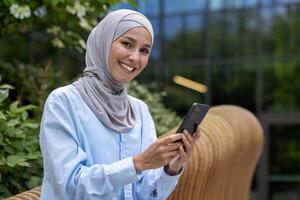 A smiling Muslim woman in hijab operates a smartphone outdoors with an office building backdrop, exuding confidence and professionalism. photo