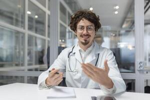 un médico vistiendo un blanco laboratorio Saco y un estetoscopio es sentado a un mesa dentro un médico oficina. sonrisa mirando cámara, hablando llamar, utilizando computadora portátil, cámara web foto