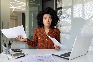 Portrait of frustrated and upset businesswoman financier, african american woman looking unsatisfied at camera, accountant worker holding reports with negative achievement results inside office. photo