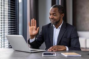 A man in a suit is sitting at a desk with a laptop and a tablet. He is waving at the camera photo