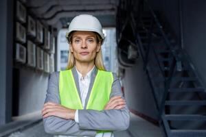 retrato de un joven mujer ingeniero, arquitecto, propietario de un construcción empresa en pie en un difícil sombrero y reflexivo chaleco fuera de el edificio y cruce su brazos, mirando seriamente dentro el cámara. foto