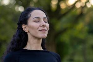 un sereno Hispano mujer cierra su ojos, disfrutando un tranquilo momento al aire libre rodeado por verdor, sensación calma y pacífico. foto