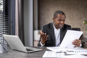 Confused male in shirt and jacket squinting in misunderstanding while reading information on paper shit. Overloaded worker getting unpleasantly surprised by amount of tasks while sitting by table. photo