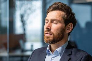 Close up businessman with closed eyes dreaming and breathing fresh clean air, mature boss in business suit thinking about future business plans and strategy, man with beard working inside office photo
