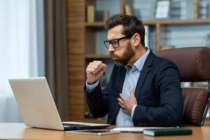 Concerned mature businessman feeling chest pain while working at his office desk, holding his chest with a strained expression. photo