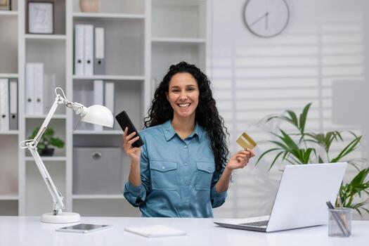 contento Hispano mujer de negocios con teléfono inteligente y crédito tarjeta, trabajando desde un acogedor moderno hogar oficina, sonriente con confianza foto