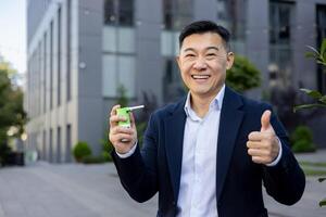 Portrait of a young Asian man in a business suit sitting on the street near the office, holding a cough spray in his hand and showing a super gesture to the camera. photo