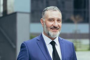 retrato de mayor hermoso empresario hombre con gris pelo y marrón pelo en negocio traje con atar. cerca el oficina centro, mira a el cámara, sonrisas foto