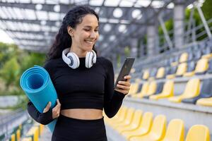 un joven atleta con yoga estera y auriculares cheques su teléfono a un vacío Deportes estadio, preparando para un ejercicio. foto