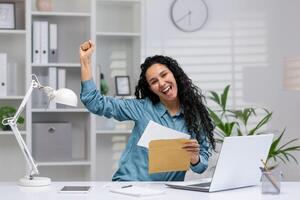 alegre Hispano mujer trabajando desde hogar celebra un negocio victoria, riendo con un elevado puño en un moderno oficina ajuste. foto