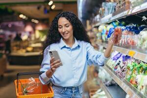 un joven mujer es en pie en un supermercado con un cesta. sostiene el teléfono, elige Cereza Tomates foto