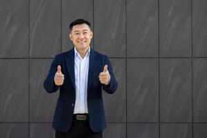 Smiling Asian businessman standing outdoors, giving two thumbs up with a modern building backdrop, conveying approval and success. photo