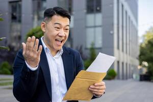 contento y sorprendido joven asiático masculino empresario en pie fuera de en el calle en un sombrero y celebrando leyendo un recibido letra en un sobre. foto