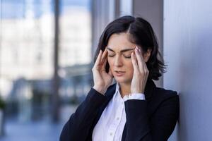 de cerca foto de un joven cansado mujer de negocios en pie en el calle y participación su cabeza, sensación dolor y presión, haciendo un masaje con su manos, cerrado su ojos.