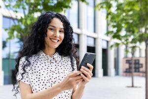 Joyful woman with curly hair, using smartphone outdoors in urban setting, exuding positivity and modern lifestyle. photo