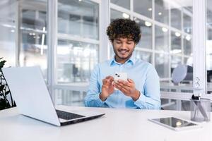 joven indio programador dentro moderno oficina sentado a lugar de trabajo, hombre utilizando teléfono, pruebas en línea solicitud en nuevo software, empresario empresario en camisa sonriente y contento con logro. foto