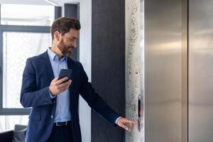 un sonriente joven hombre empresario es en pie en un oficina espacio cerca el ascensor y prensas un llamada botón, participación un teléfono en su manos. foto