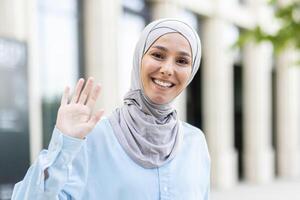 Portrait of a modern Muslim woman wearing a hijab, greeting with her hand raised, standing outdoors in a sunny environment. photo