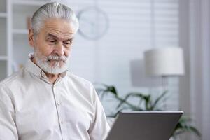 An elderly gentleman focuses intently on a digital tablet while seated in a well-lit, contemporary room, suggesting technology use among seniors. photo