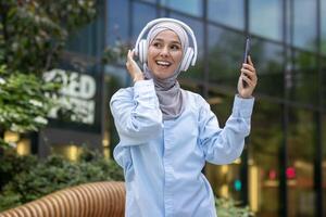 A cheerful woman in a hijab listens to music with headphones and dances joyfully outside an office building. photo