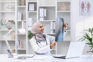 sonriente musulmán hembra médico en hijab revisando un radiografía en un bien iluminado médico oficina, reflejando cuidado de la salud profesionalismo y diversidad. foto