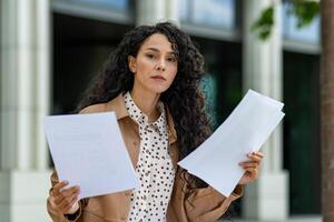 A focused Hispanic businesswoman holding and reviewing multiple documents outside a modern office building. She appears confident and professional. photo