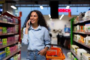 sonriente Hispano mujer comprador en supermercado, camina Entre filas de estantería con bienes, mujer alegremente negociaciones en móvil teléfono, comprador recomienda Tienda a amigo foto