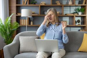 Elderly remote worker holding portable computer on laps while taking off glasses and making break for rest. Businesswoman rubbing nose for relieving tired eyes from long time looking at screen. photo