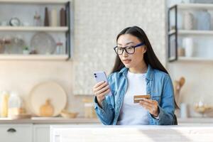 financiero problemas. preocupado joven asiático mujer participación teléfono y crédito tarjeta. sentado en el cocina a hogar, molesto a resolver un problema. foto