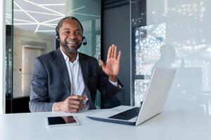 retrato de un sonriente joven africano americano hombre en un traje sentado a un escritorio en un auriculares y en frente de un computadora portátil, ondulación y saludo a el cámara. foto