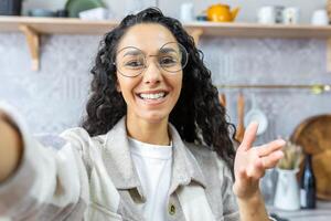 Close up photo. Young beautiful Latin American woman talking on call at home, blogging, taking selfie, smiling. Waving at camera. photo