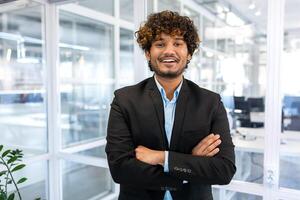 Portrait of successful hispanic boss, businessman in business suit looking at camera and smiling, man with crossed arms working inside modern office building. photo