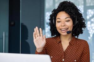 Woman with headset phone using laptop for remote communication and call, african american woman waving at camera greeting. photo