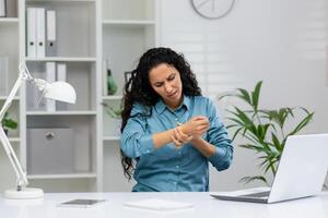 un adulto mujer en un azul camisa siente muñeca dolor mientras trabajando a su oficina escritorio, demostración incomodidad y cepa. foto