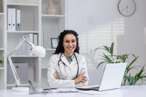 A cheerful Hispanic doctor sits confidently at her desk with a laptop, showcasing professionalism and approachability. photo