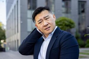 Close-up photo of a tired young Asian man sitting outside in a suit and holding something with his hand, giving himself a massage from pain and tension.